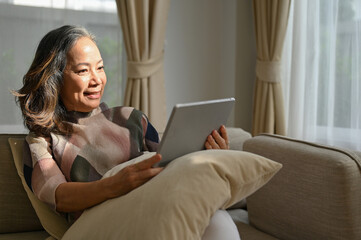 Fototapeta na wymiar Attractive asian retired woman relaxes in the living room and using portable tablet.