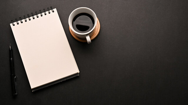 Black workspace table with a cup of coffee, an empty spiral notepad and copy space
