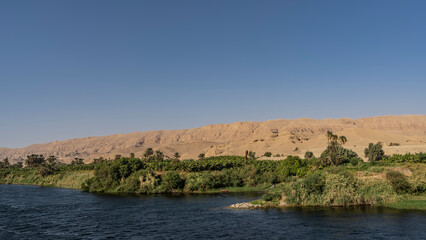 The Nile River flows calmly. Ripples on the blue water. There is green vegetation on the shore. Sand dunes against a clear sky. Egypt.