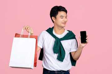Smiling young handsome southeast Asian man holding mobile phone and shopping bags in pink studio...