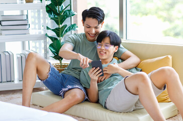 Two Asian young handsome pride male gay men lover couple partner sitting smiling laying down together on cozy sofa in bedroom cuddling hugging watching entertainment media streaming on smartphone