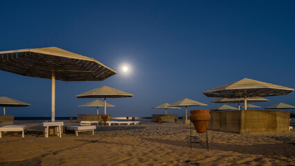 A quiet night on the Red Sea beach. The full moon is shining in the dark blue sky. Lattice umbrellas, empty sunbeds, urns are on the sand. Egypt. Safaga
