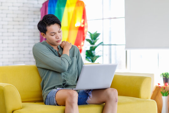 Asian young teenager male gay businessman sitting on sofa holding laptop notebook computer working online from home in living room with rainbow equality unity flag celebrate LGBTQ June pride month