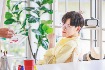 Asian young handsome teenager gay man model in casual outfit sitting posing gesture while unrecognizable painter using paintbrush drawing painting color picture on canvas on wooden easel in studio