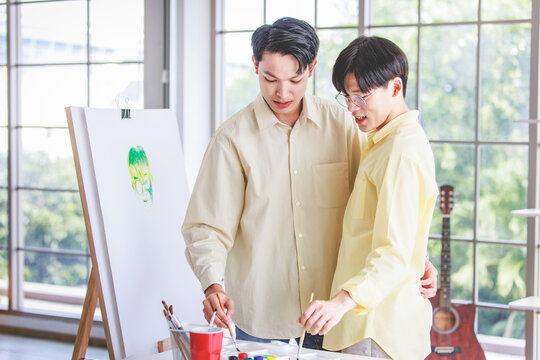 Two Asian Young Lovely Pride Teenager Male Gay Men Lover Couple Partner Standing Smiling Having Fun Playing Paintbrush In Front Canvas Painting Board On Easel In Glass Windows Workshop Studio At Home
