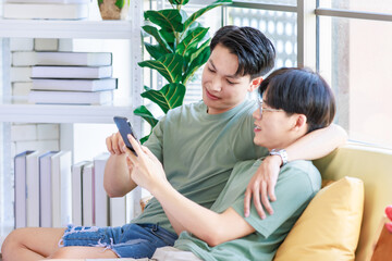 Two Asian young handsome pride male gay men lover couple partner sitting smiling together on cozy sofa in bedroom holding looking browsing surfing via smartphone cuddling hugging showing LGBTQ love