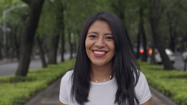 Portrait of a cute latino woman with beautiful eyes and tanned skin looking at camera while laughing out loud on an avenue with blurred traffic lights and trees, medium shot