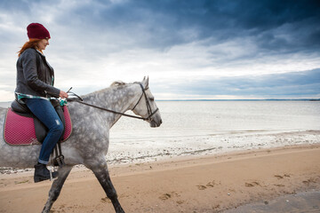 wearing jeans, warm hat and jacket horsewoman rides astraddle along sandy beach