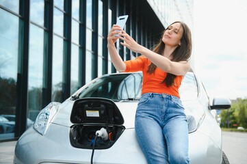 Young woman is standing near the electric car and looks at the smart phone. The rental car is charging at the charging station for electric vehicles. Car sharing.