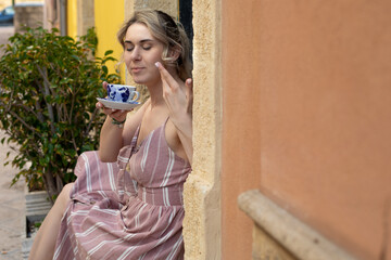 Fototapeta premium Portrait of young woman with fair hair wearing summer pink striped dress, sitting on step, enjoying aroma of coffee.