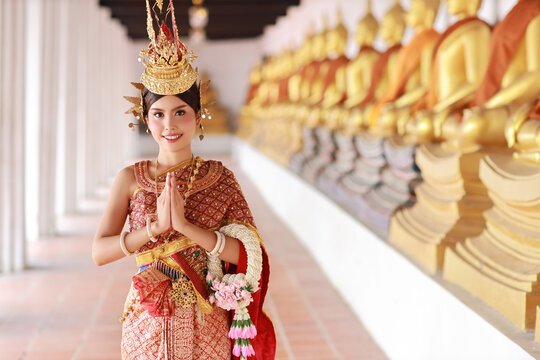 Young fashion and beautiful asian woman in red Thai traditional costume pay respect sawasdee symbol while holding jasmine garland standing in ancient buddha temple Ayutthaya, Thailand. Travel concept