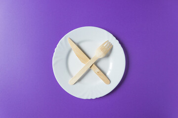 Wooden fork and knife lie on a white plate crosswise. Tableware from natural materials isolated on a purple background.
