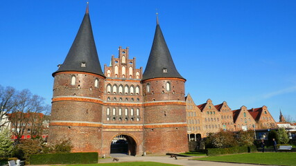 Obraz premium Lübeck Holstentor mit zwei Spitztürmen neben Salzspeichern und Grünanlage unter blauem Himmel