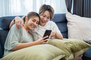 Happy two lesbian woman, lgbtq homosexual Lesbian couples sharing earphones while listening music over smart phone in living room. Gay woman couple rest and relaxing by using smartphone in bedroom. 