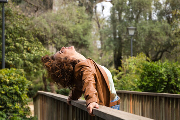 Beautiful, curly-haired woman leaning on the railing of a wooden bridge with her head tilted back....