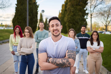 Man with tattooed arms smiling looking at the camera while standing in front of a group of people . Team concept and leadership.