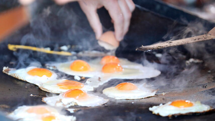 A number of fresh raw eggs sizzling away on a smoking hot black bbq cook top. An egg is cracked in the background to be cooked. Eggs on the barbecue sunny side up yolks.