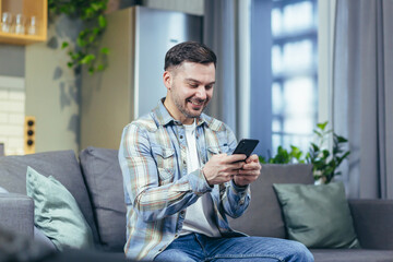 The happy man looks at the phone at home and rejoices in success and victory