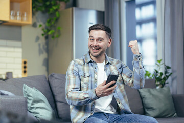 The happy man looks at the phone at home and rejoices in success and victory