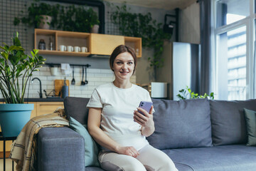 Obraz premium Happy pregnant woman at home rejoices with phone smiling at home and looking at camera