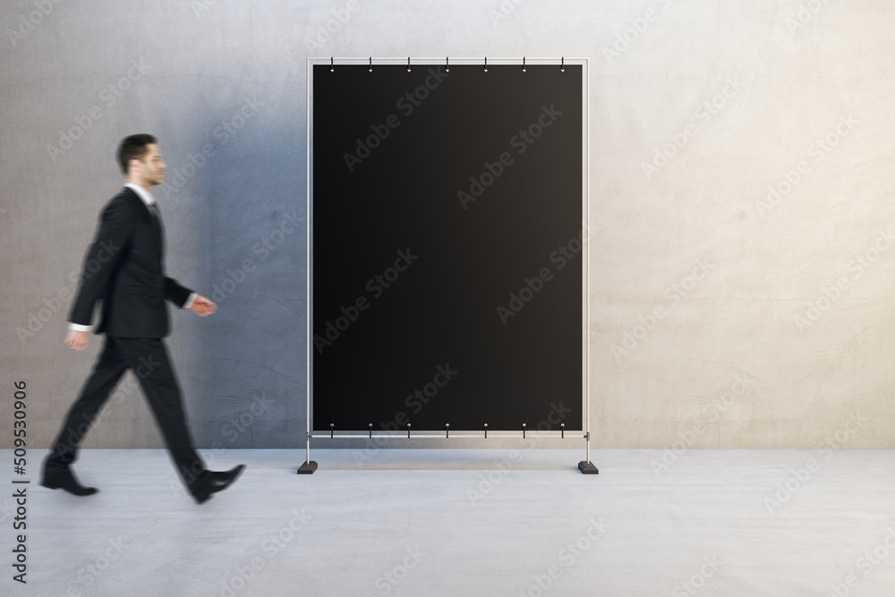 Poster Side view of attractive young man walking past empty black rectangular poster in simple concrete interior. Advertisement and mock up concept.
