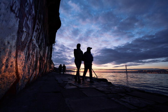 Silhouette of walking couple on river Tagus bank in Lisbon at sunset time.