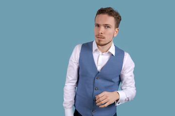 What does not hear you. Handsome business guy in white shirt and blue vest, holding his hand next to his ear and leaning over with a question, a caucasian young man posing on a blue background