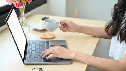 Cropped shot young woman holding coffee cup and using computer laptop in modern home kitchen
