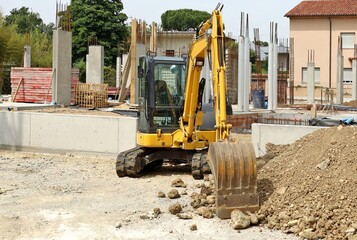 Compact excavator at work with the foundations and the pillars of a residential building under construction  behind it.