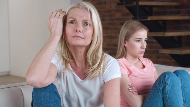 Senior Mother and her young adult daughter turn back on sofa after conflict. Resentful daughter and mature mother feel upset having misunderstanding.