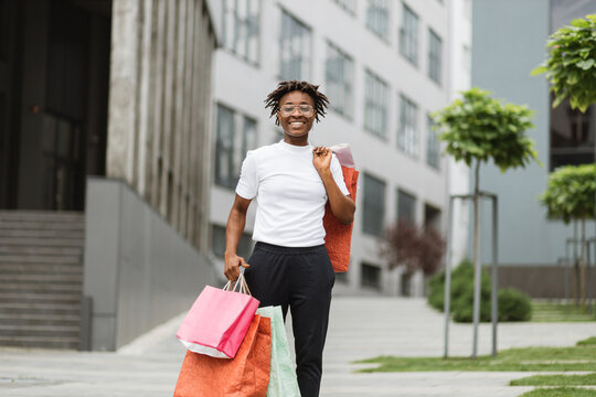 Shopping, urban lifestyle concept. Horizontal shot of happy young smiling black woman with curly hair, posing to camera with colorfull shopping bags against beautiful modern city background.