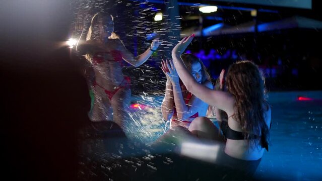 Cheerful young people splashing water to girls in swimwear, dancing and partying in luxury resort. Friends have night pool party in a private villa swimming pool. Women hanging out. Slow motion.