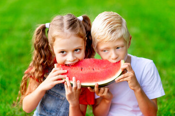 children girl and boy blonde in summer on the lawn with eating watermelon on the green grass having fun and rejoicing biting it, space for text