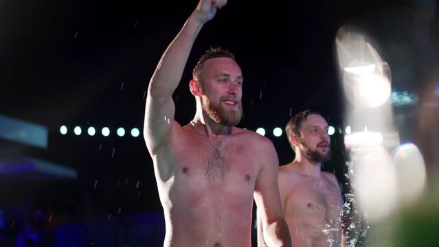 Happy homosexual man celebrating gay pride with friends at night LGBTQ pool party in a private villa swimming pool. Bisexual male in swimwear dancing splashing water in luxury resort. Slow motion.