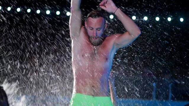 Happy homosexual man celebrating gay pride with friends at night LGBTQ pool party in a private villa swimming pool. Bisexual male in swimwear dancing splashing water in luxury resort. Slow motion.