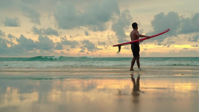 4K Healthy Asian senior man surfer in swimwear holding surfboard walking on the beach at summer sunset. Elderly retired male enjoy outdoor activity lifestyle and water sport surfing on summer vacation