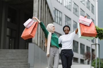 City shopping concept. Outdoor urban lifestyle portrait of two diverse young women, African and Muslim, holding colorful shopping bags while walking in the modern city.