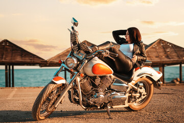 Cool confident adult woman in leather jacket and boots posing lying on motorcycle. Beach umbrellas...