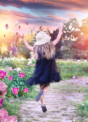 Fototapeta premium A little girl in a black airy dress and hat jumps with joy in the botanical garden where peonies bloom. Happiness concept