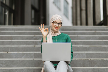 Smiling young woman in hijab woking on wireless laptop while sitting on stairs near modern...