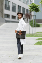 Full length portrait of young attractive African woman doctor healthcare worker in white coat, standing with clipboard folder in hands outdoors in front of modern hospital or clinic