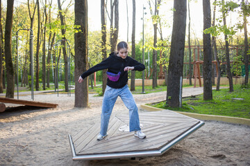 child walks on playground. girl balances on simulator against background of wooden rides and trees in nature park