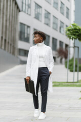 Portrait of young smart focused african female medical intern doctor with folder clipboard, standing outside modern hospital building. Medicine, education and healthcare concept.