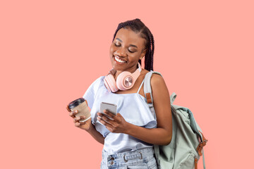 Cheery young black woman with backpack, headphones and takeaway coffee using cellphone on pink studio background