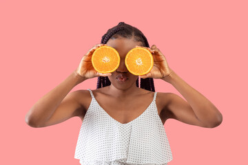 Young black woman with braids holding orange halves near her eyes, puckering lips for kiss on pink studio background
