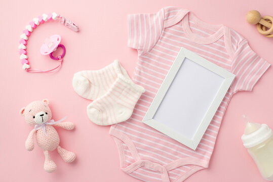 Baby concept. Top view photo of photo frame pink infant clothes bodysuit socks baby's dummy chain bottle wooden rattle and knitted teddy-bear toy on isolated pastel pink background with empty space
