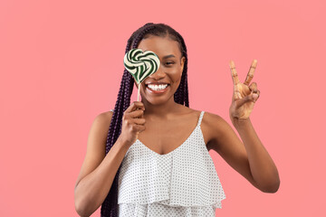 Joyful young black woman holding heart shaped candy on stick, covering one eye with lollipop,...