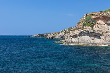 Fototapeta na wymiar Rocky coast of Ibiza island scorched by the summer sun with sparse vegetation, calm peaceful sea, clear day, not a cloud in the sky, Balearic Islands, Spain
