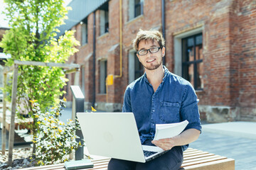 Portrait of a young teacher outside the university campus, sitting on a bench with a laptop and smiling