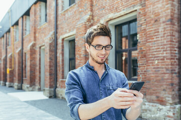 Fototapeta premium Portrait of young male student, outside campus holding cellphone, smiling and walking